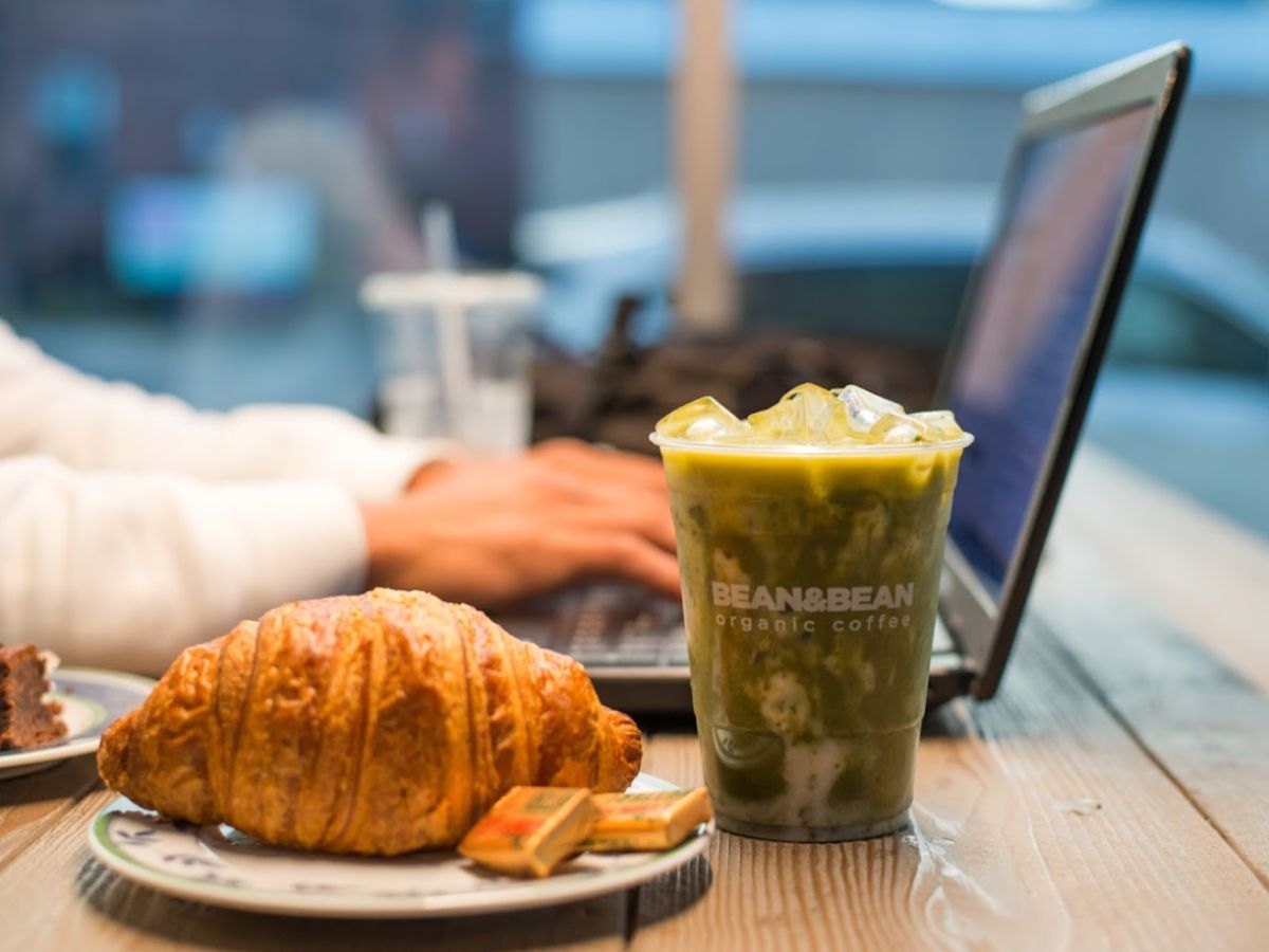 A croissant and a Bean & Bean iced matcha latte on a table. In the background, a person works on their laptop. 