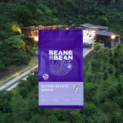 Purple Bean & Bean coffee bag labeled “Altieri Estate Gesha” displayed in front of a lush hillside coffee farm with the Altieri Specialty Coffee facility in the background.