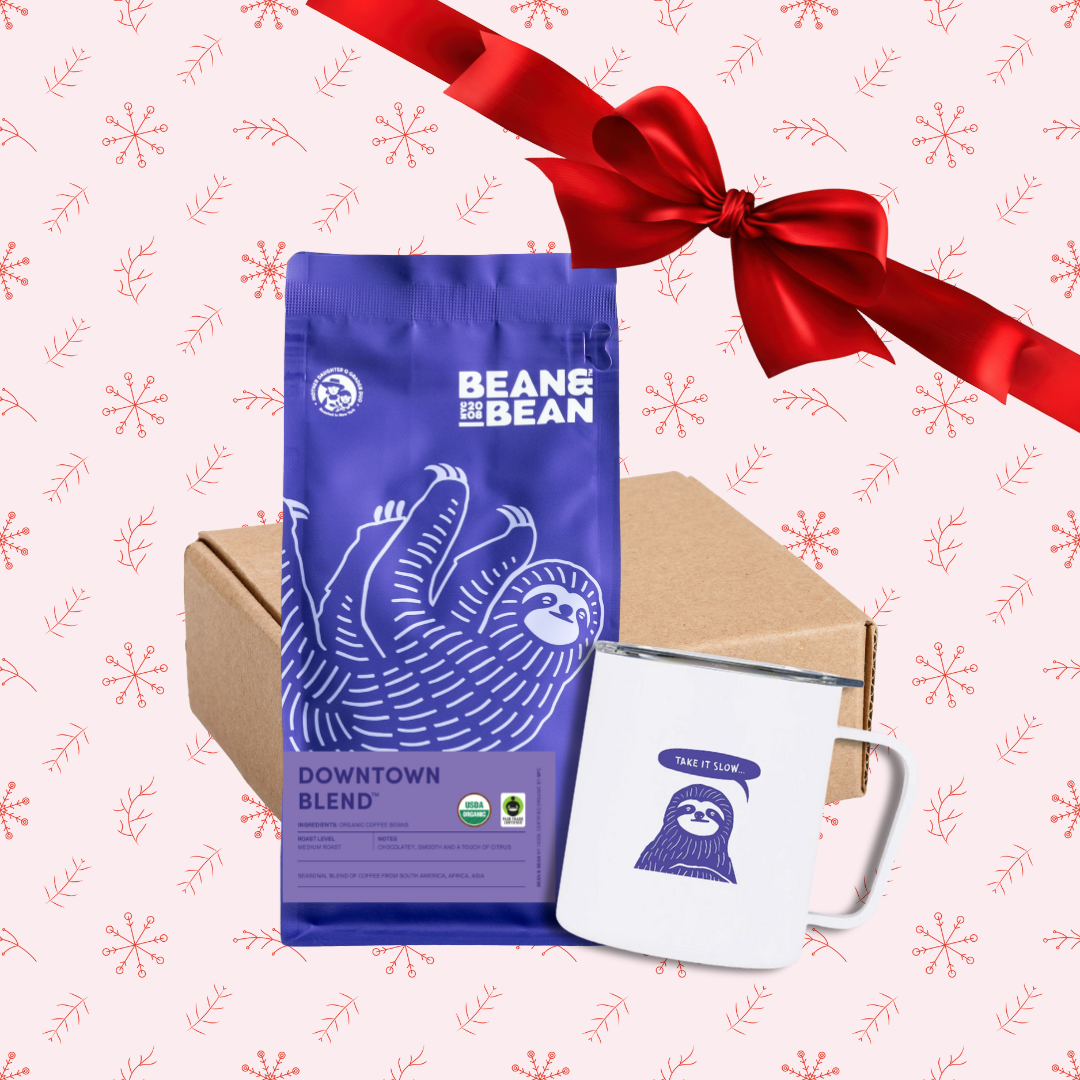 Gift set with a purple Bean & Bean Downtown Blend coffee bag, a white camp mug with a sloth illustration, and a kraft box, displayed on a pink background with red snowflake patterns and a red ribbon.