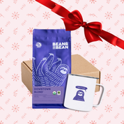 Gift set with a purple Bean & Bean Downtown Blend coffee bag, a white camp mug with a sloth illustration, and a kraft box, displayed on a pink background with red snowflake patterns and a red ribbon.