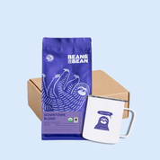 Gift set with a purple Bean & Bean Downtown Blend coffee bag, a white camp mug with a sloth illustration, and a kraft box, displayed on a blue background 