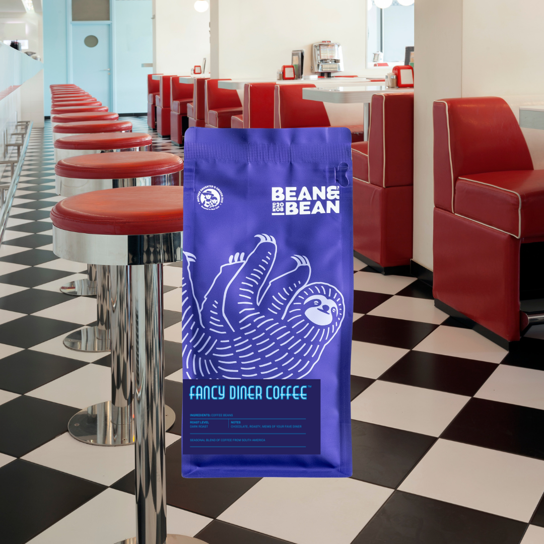 A purple Bean & Bean coffee bag with a large white sloth illustration, placed in front of a retro diner interior with red booths, chrome stools, and a black-and-white checkered floor.