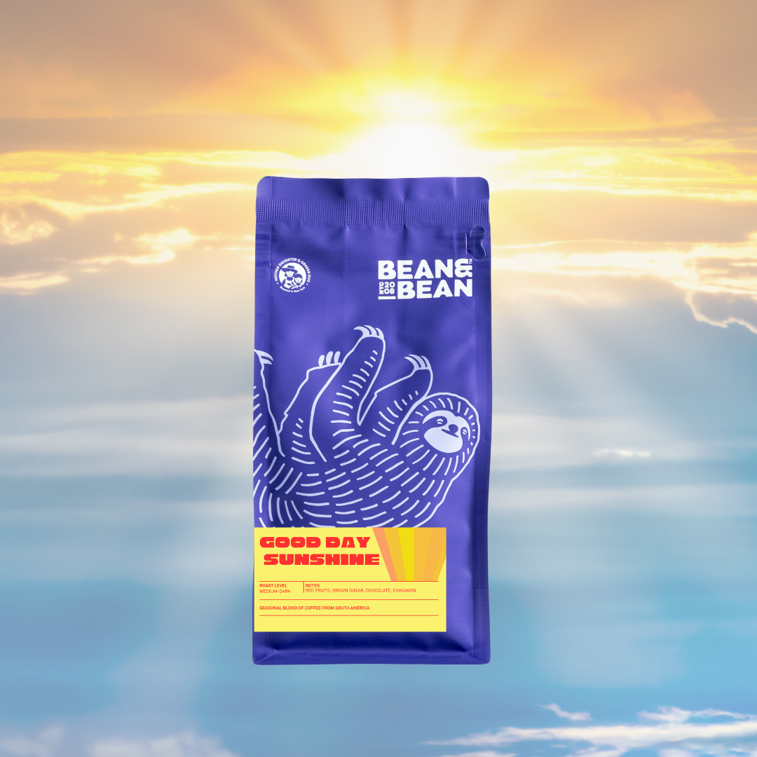 A purple Bean & Bean coffee bag with a large white sloth illustration and a yellow “Good Day Sunshine” label, set against a bright sky background with sun rays breaking through clouds.