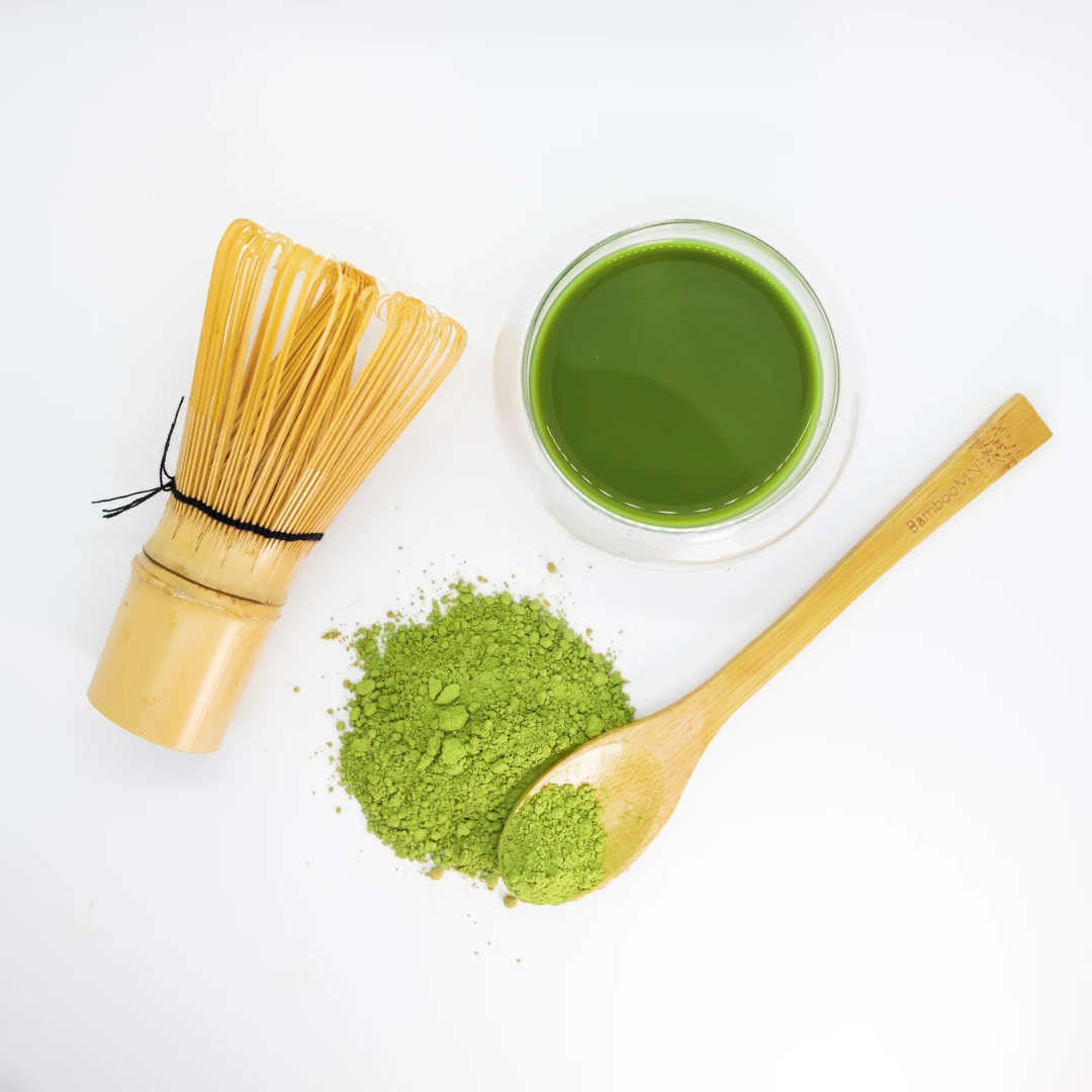 A bamboo matcha whisk, a glass of prepared matcha usucha, a wooden spoon, and a small pile of bright green matcha powder.