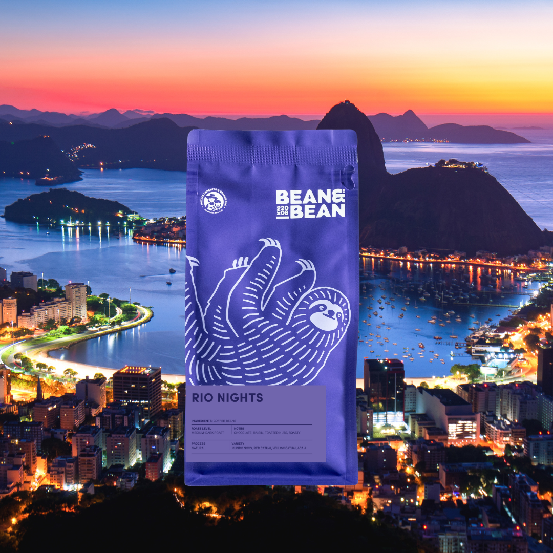 A purple Bean & Bean coffee bag with a white sloth illustration and a “Rio Nights” label, shown in front of a vibrant coastal city and bay at dusk.