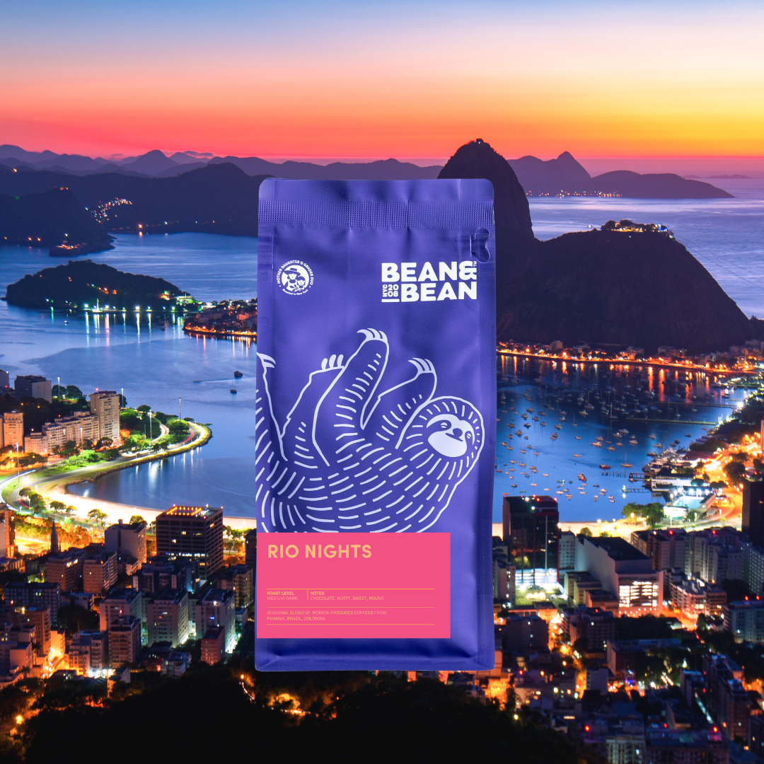 A purple Bean & Bean coffee bag with a white sloth illustration and a “Rio Nights” label, shown in front of a vibrant coastal city and bay at dusk.