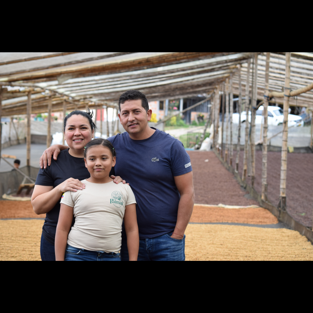 A smiling family of three stands together inside a coffee drying station, surrounded by drying coffee beans on raised beds.