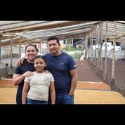 A smiling family of three stands together inside a coffee drying station, surrounded by drying coffee beans on raised beds.