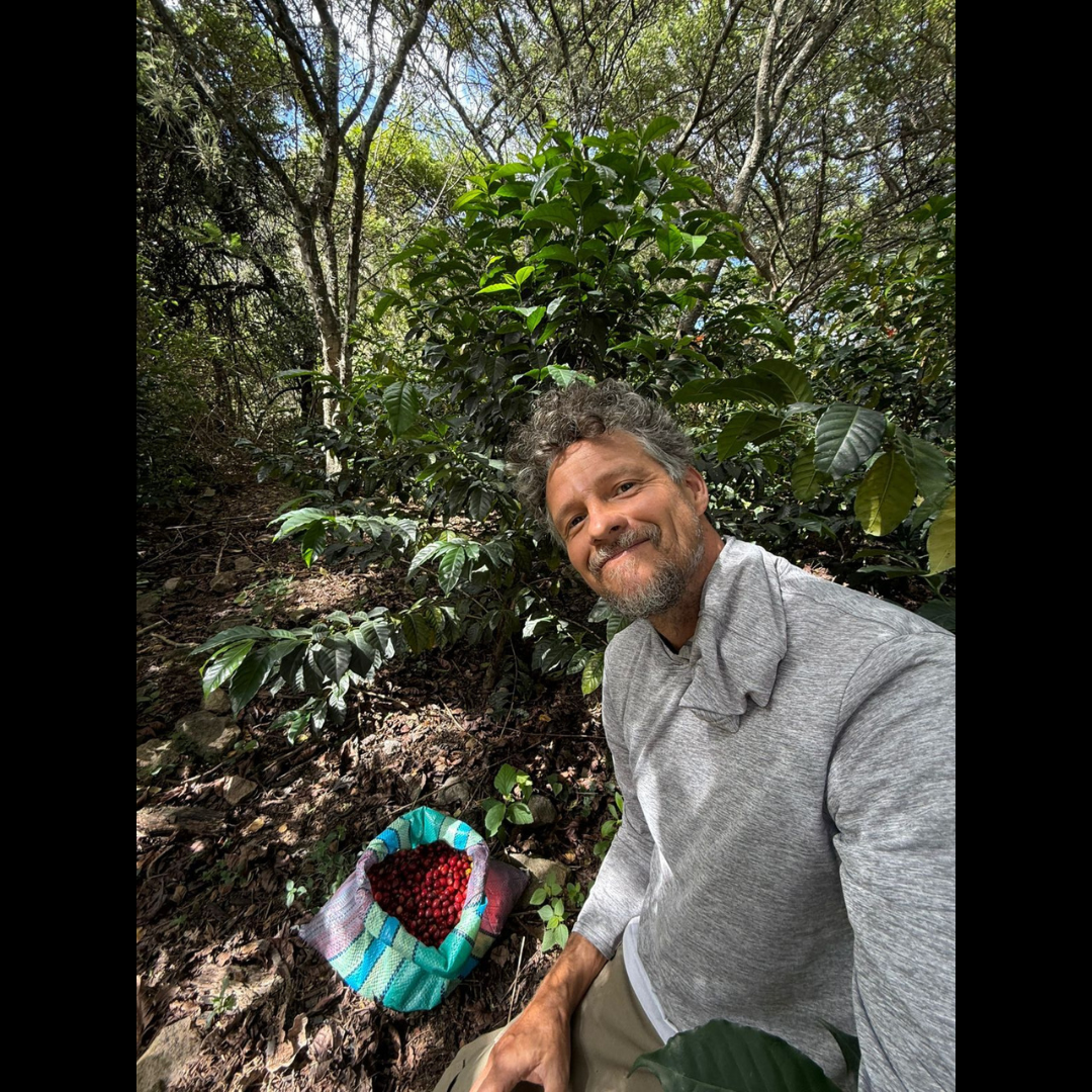 A man in a gray shirt smiling at the camera, kneeling beside a woven bag full of freshly picked red coffee cherries, with coffee plants and trees in the background.