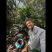 A man in a gray shirt smiling at the camera, kneeling beside a woven bag full of freshly picked red coffee cherries, with coffee plants and trees in the background.
