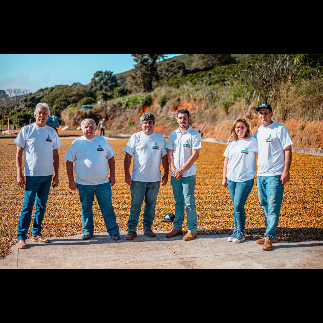 Six coffee producers in matching white farm t-shirts stand together on a patio of drying coffee cherries, with green hillsides in the background.