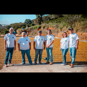 Six coffee producers in matching white farm t-shirts stand together on a patio of drying coffee cherries, with green hillsides in the background.